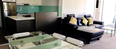Un área de estar moderna con una mesita de centro de vidrio, un sofá de cuero negro con cojines estampados, y una cocina con alacenas blancas y salpicadero verde.