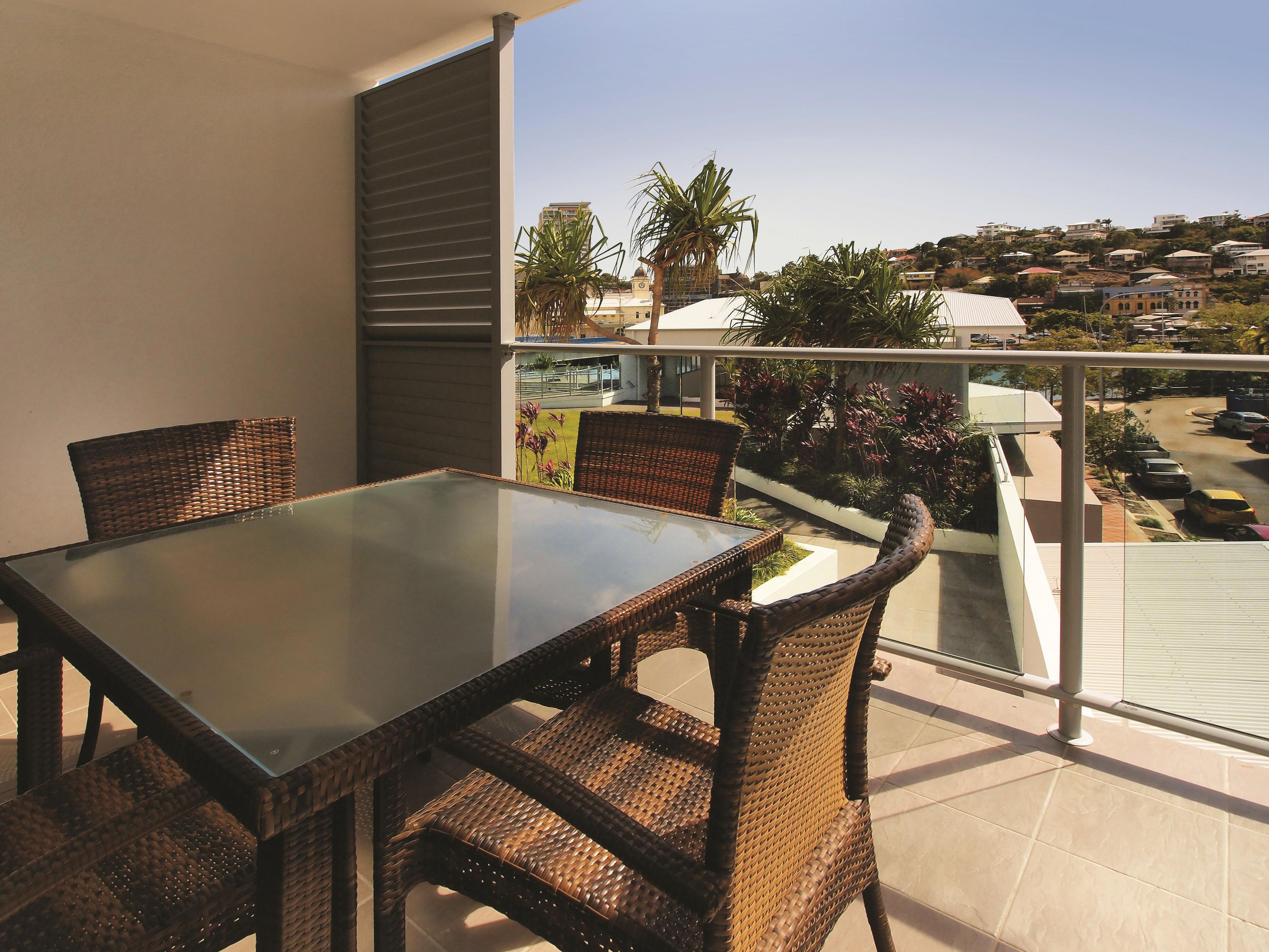 Een balkon met een glazen tafel en rieten stoelen, met uitzicht op een woongebied met huizen en geparkeerde auto's.