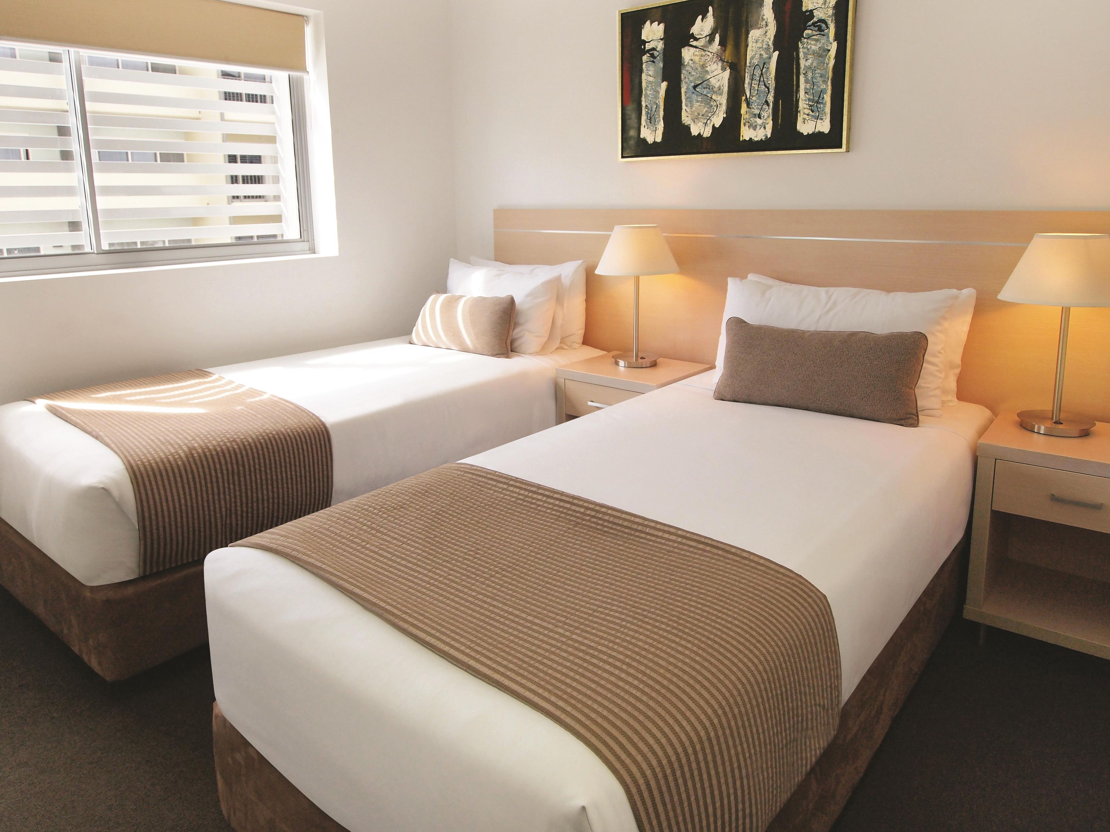 A hotel room with two beds, a window with blinds, a bedside table with a lamp, and a piece of wall art.
