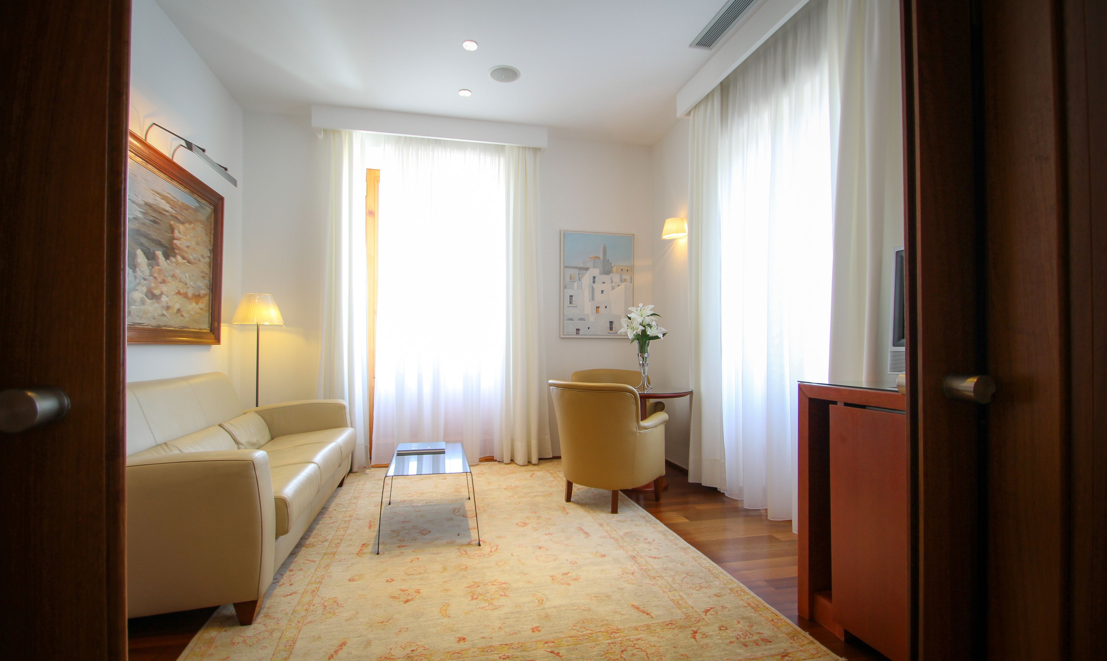 A hotel room with a sofa, a small table, a chair, a cabinet, and a painting on the wall.