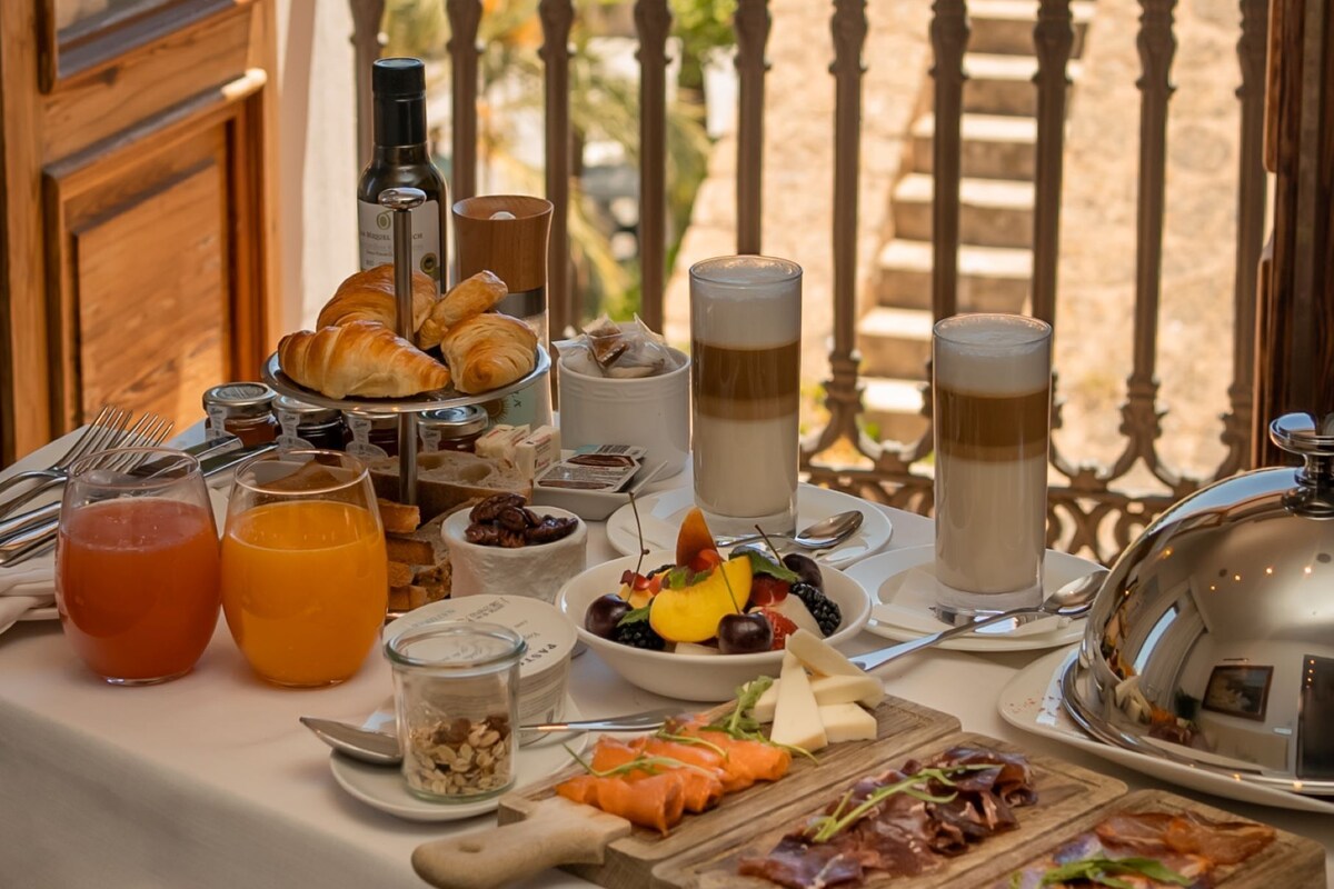 A breakfast table with croissants, fruits, and beverages, overlooking a scenic view of the sea and palm trees.