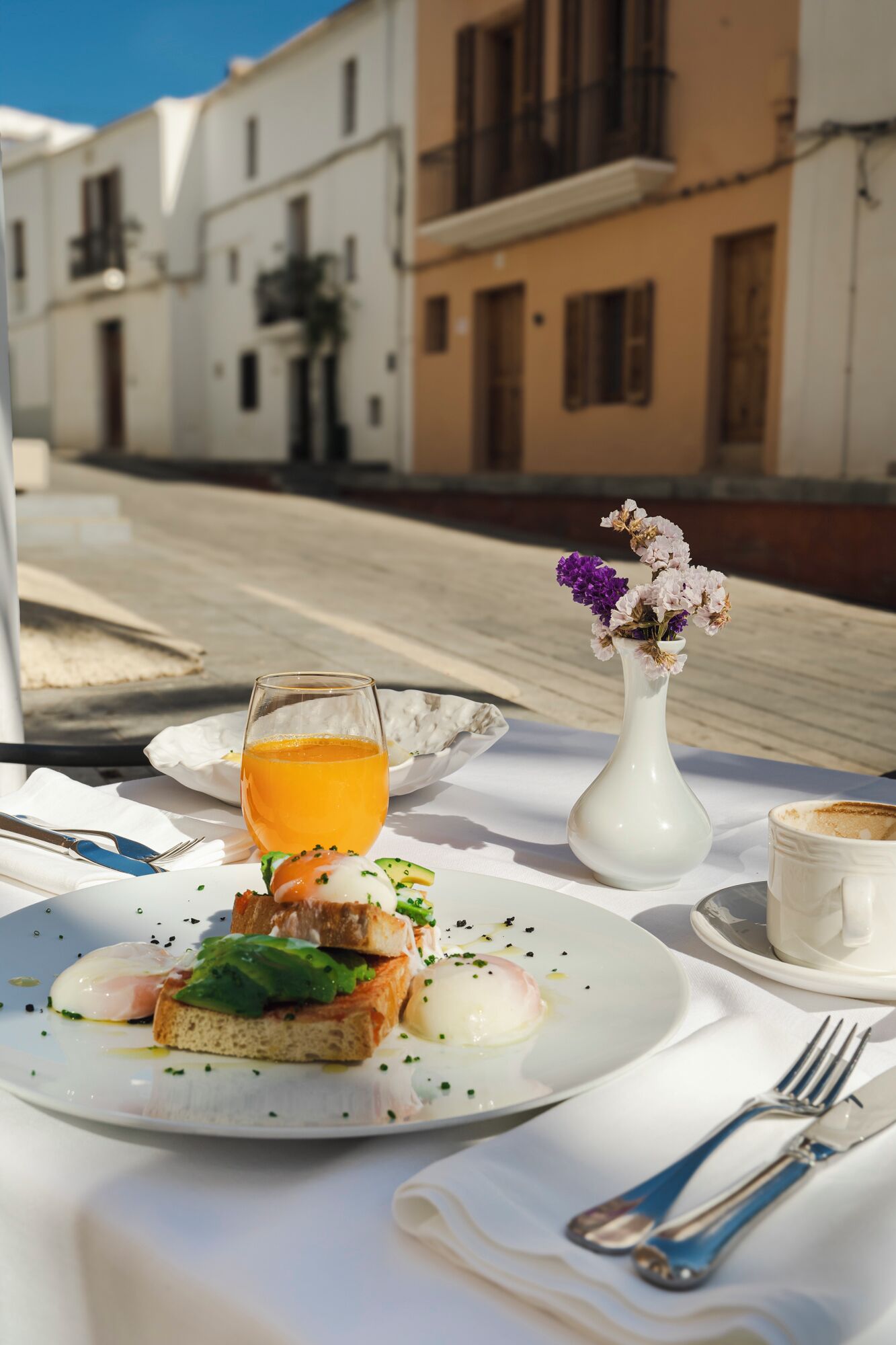 Una escena de desayuno con un plato de comida, un vaso de jugo de naranja, un jarrón blanco con flores y una taza de café sobre una mesa al aire libre.