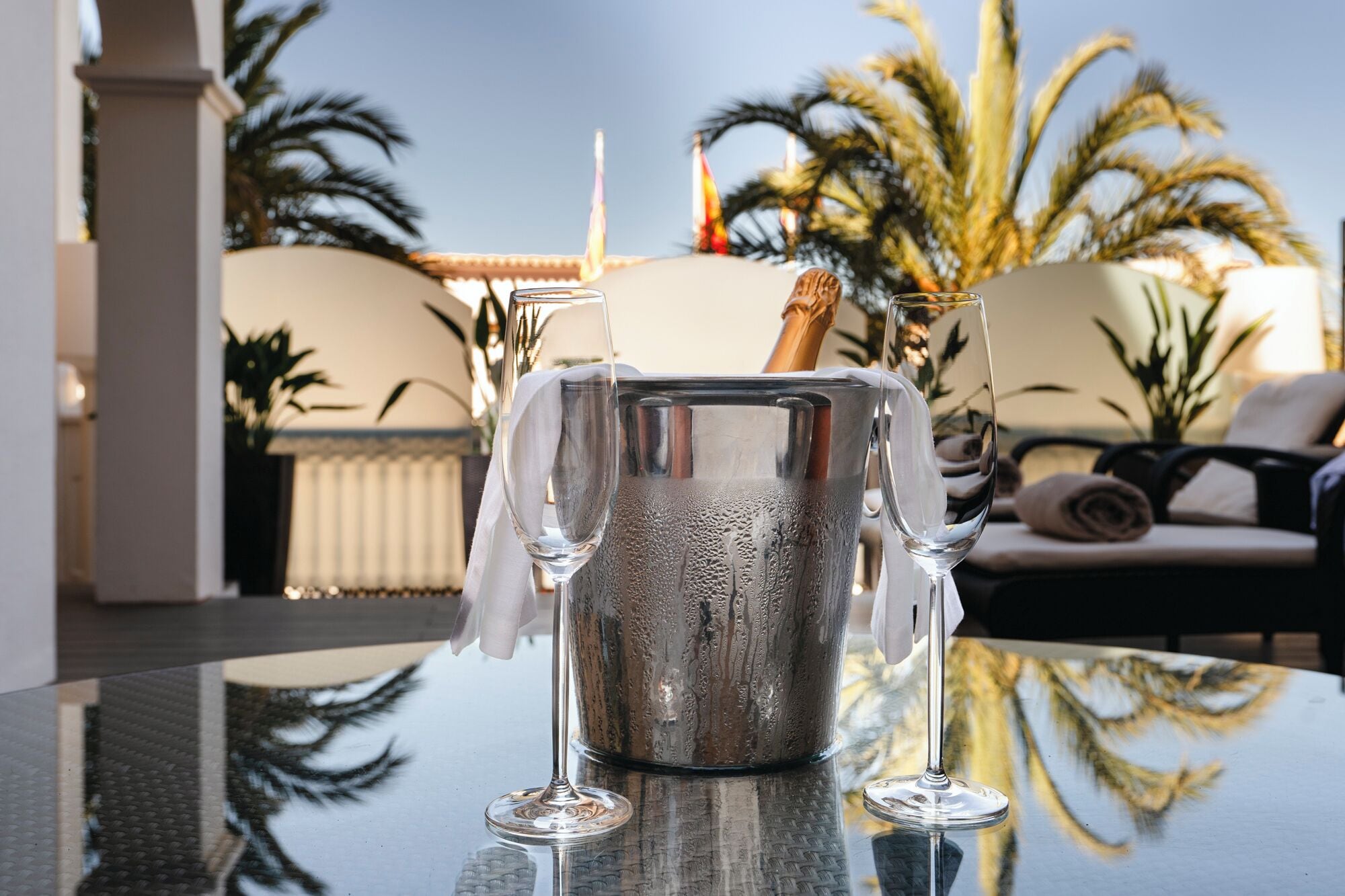 Una copa de champán sobre una mesa reflectante, con vista a palmeras y un rincón de estar acogedor al fondo.