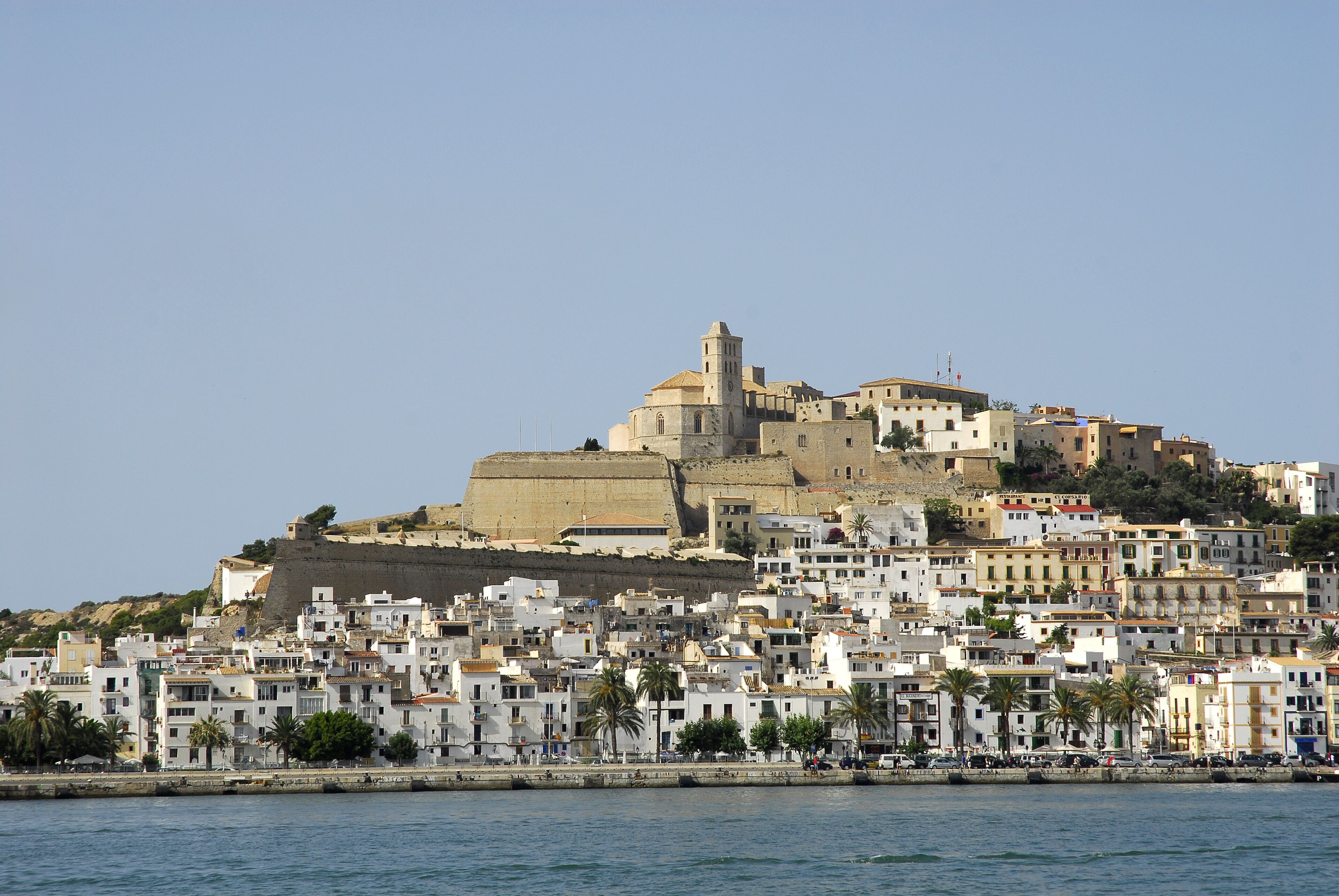 A coastal cityscape with a prominent historical fortress on a hill, surrounded by densely packed buildings with balconies and palm trees.