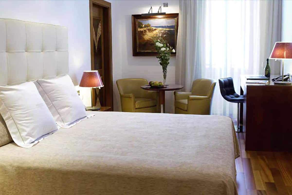 A hotel room with a large bed, two chairs, a small table with fruit, a desk, and a painting on the wall.