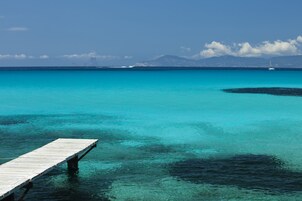 A wooden pier extending into clear turquoise waters with a distant mountain range and a sailboat in the background.