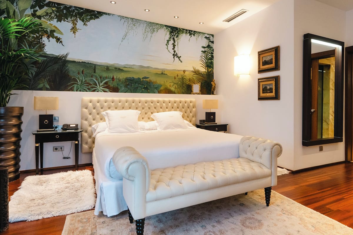 A bedroom with a large bed, two bedside tables, a mirror, and a mural on the wall.