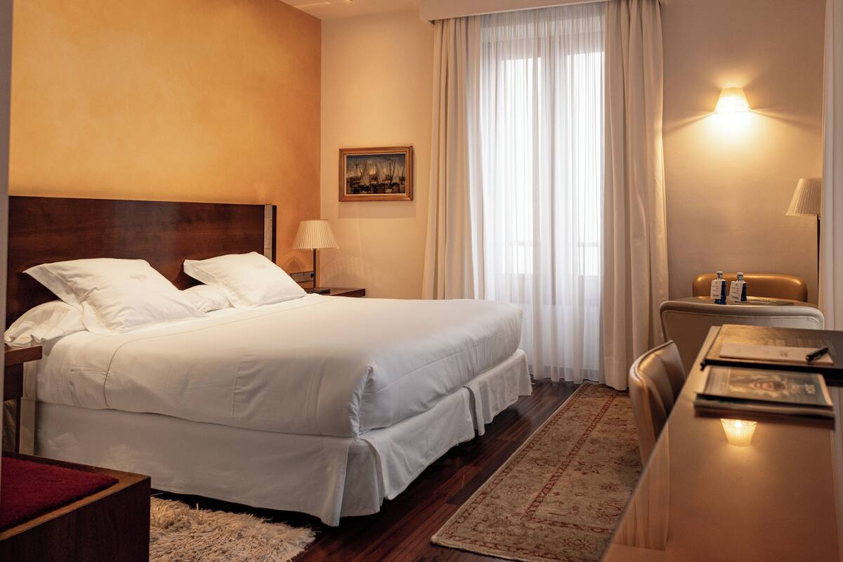 A hotel room with a large bed, a desk, a chair, a nightstand, a lamp, a painting on the wall, and a window with curtains.