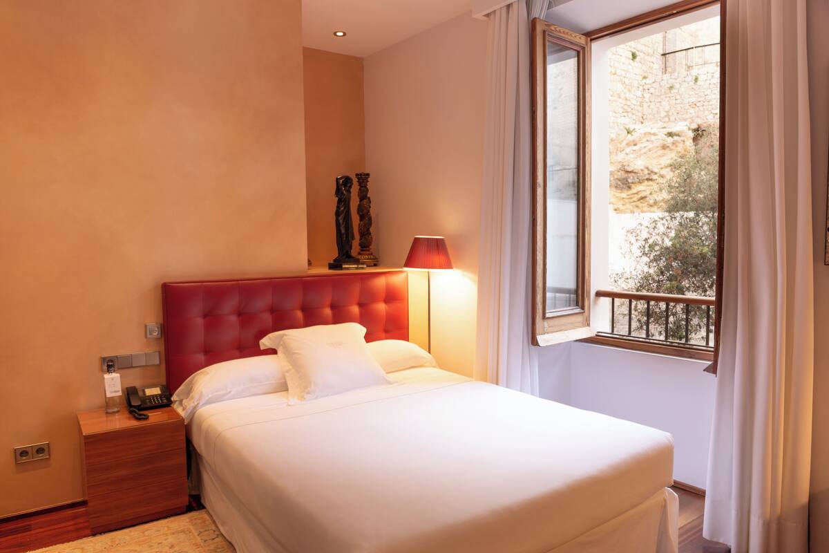 A hotel room with a large bed, a nightstand with a telephone, a red lamp, and a window with a view of a stone wall and greenery.