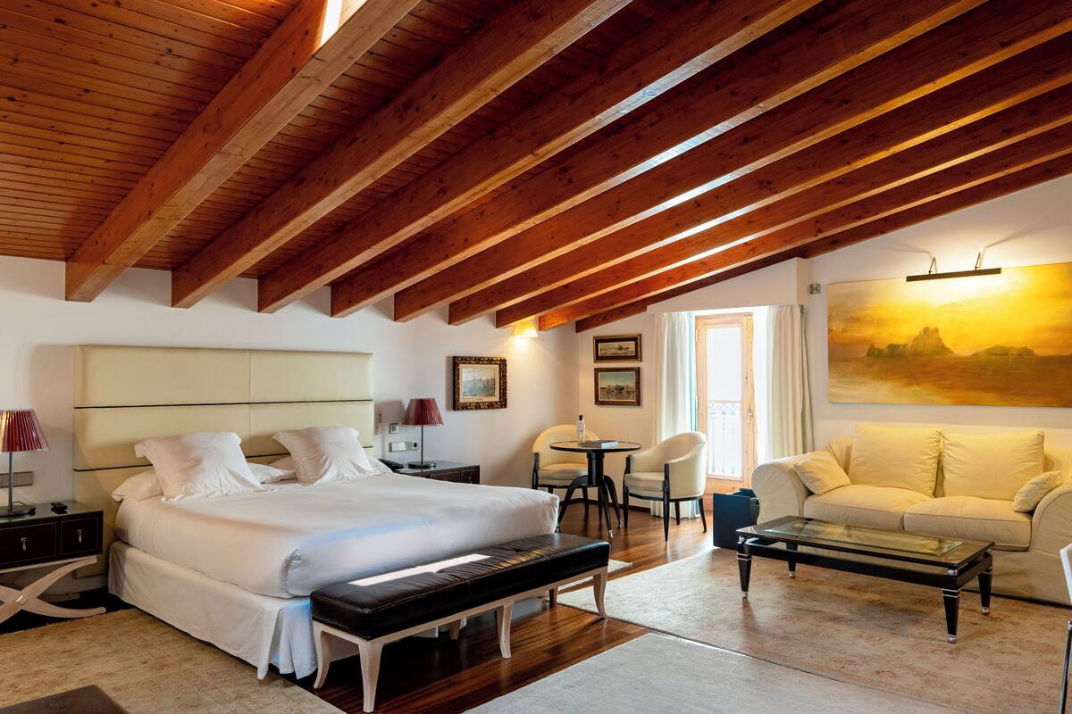 A spacious bedroom with a large bed, a bedside table, a chair, a sofa, and a painting on the wall.