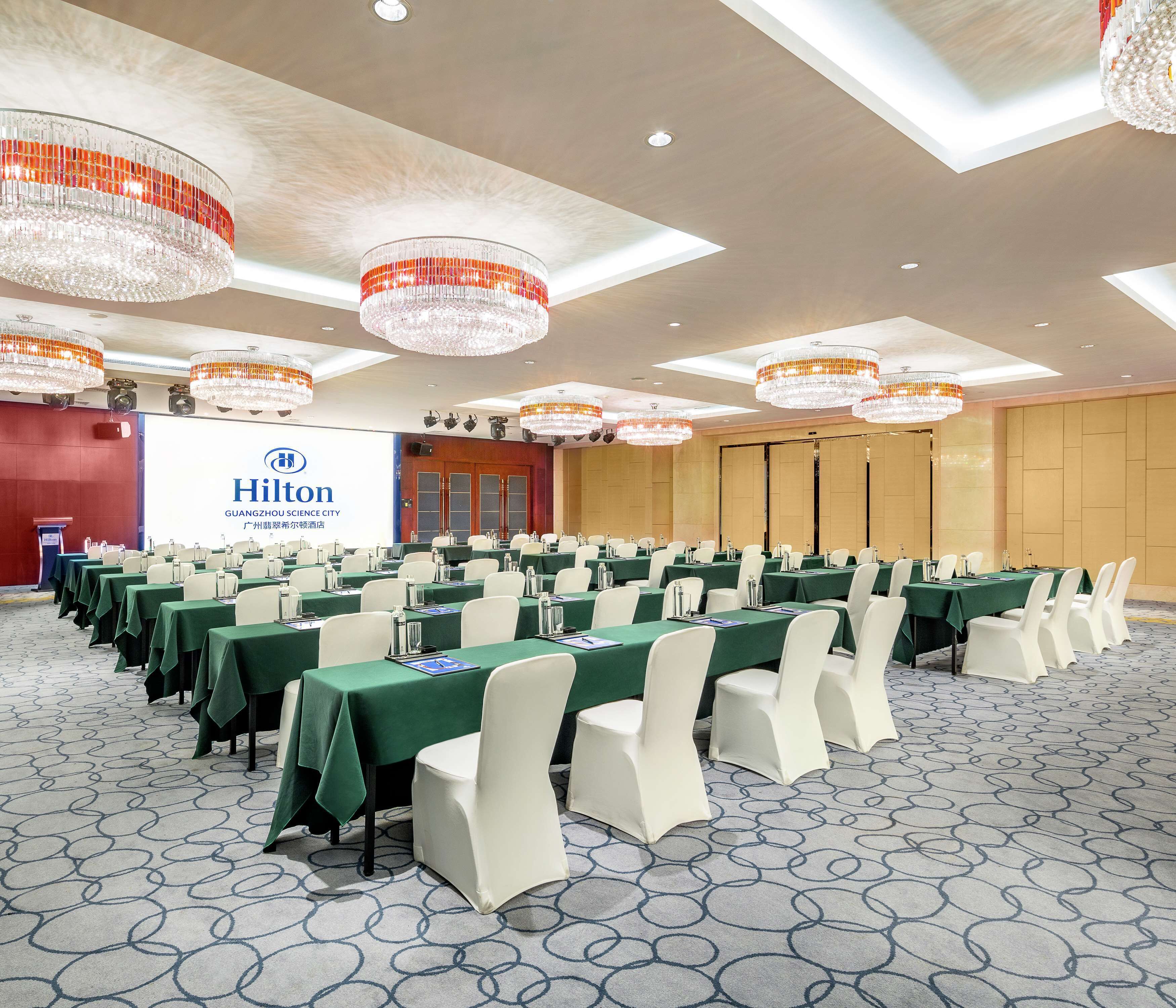 A conference room with rows of tables and chairs, a large screen displaying the Hilton logo, and chandeliers on the ceiling.