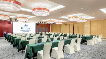 A conference room with rows of tables and chairs, a large screen displaying the Hilton logo, and chandeliers on the ceiling.