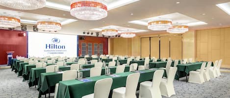 A conference room with rows of tables and chairs, a large screen displaying the Hilton logo, and chandeliers on the ceiling.