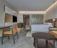 A modern hotel room with a bed, a desk, a chair, a round table, and a TV.