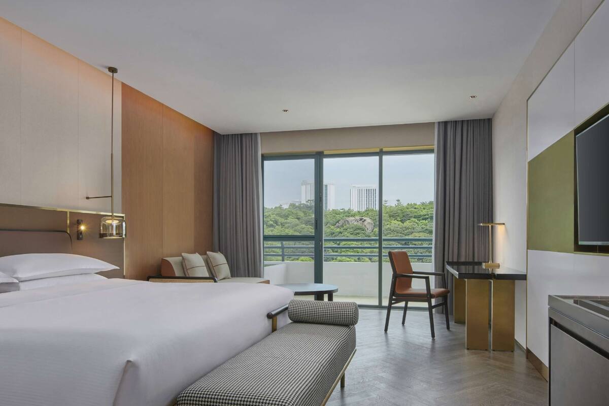 Une chambre d’hôtel avec un grand lit, un bureau, une chaise et un banc. Il y a une fenêtre donnant sur des bâtiments et de la verdure.