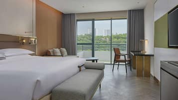 Ein Hotelzimmer mit einem großen Bett, einem Schreibtisch, einem Sessel und einer Bank. Ein Fenster bietet Blick auf Gebäude und Grünflächen.