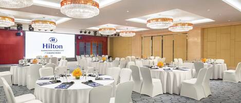A spacious conference room with multiple round tables, white chairs, and elegant chandeliers.