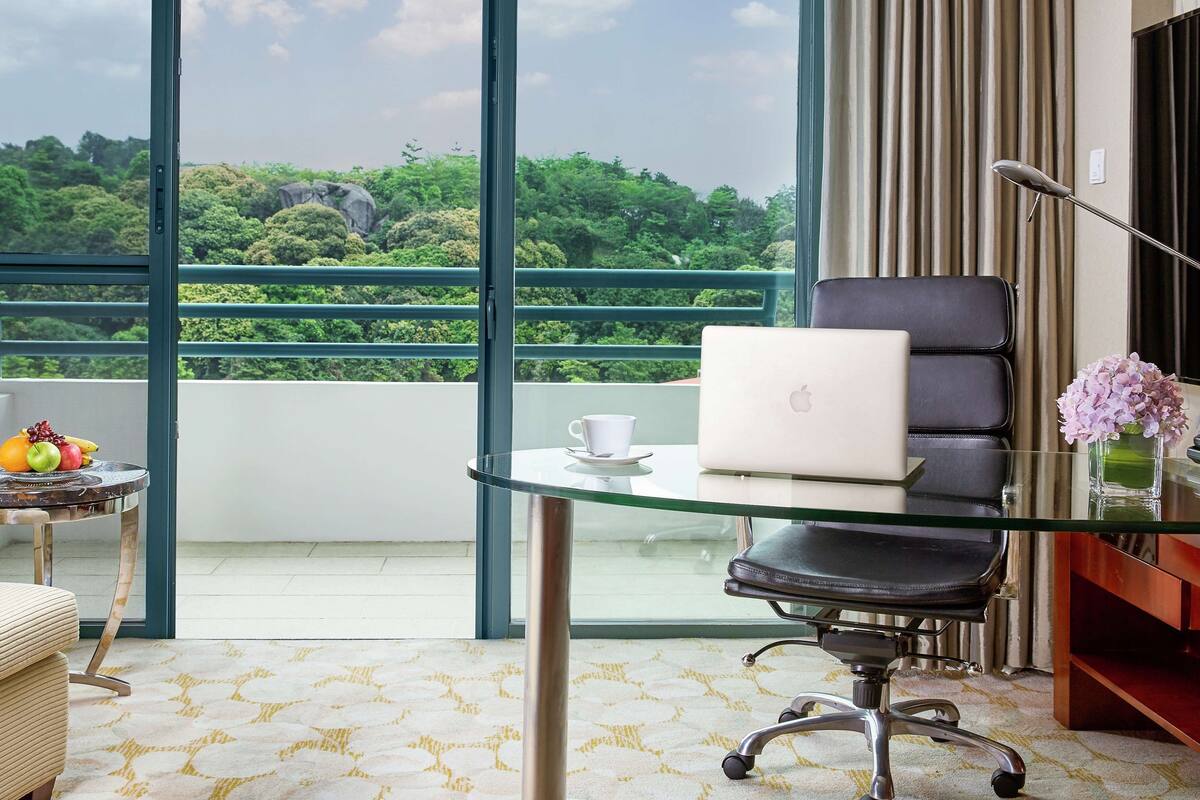Une chambre d’hôtel moderne dotée d’un grand balcon, d’une table en verre avec un ordinateur portable, d’une chaise et d’une petite table sur laquelle se trouvent des fruits et une tasse.