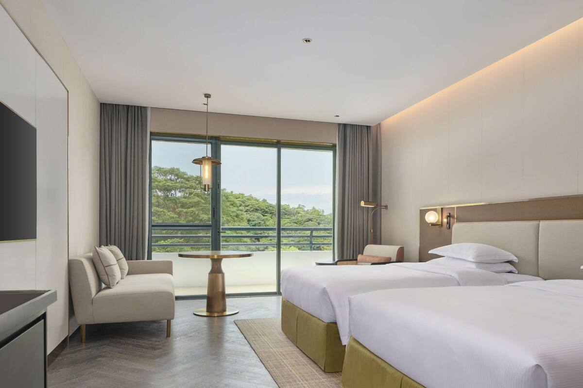 Une chambre d’hôtel avec deux lits, un canapé, une petite table et une grande fenêtre donnant sur un paysage verdoyant.