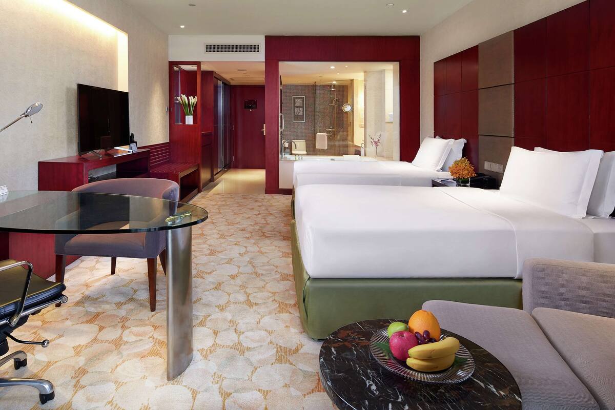 Une chambre d’hôtel avec deux lits, un bureau avec une télévision, une table en verre et un panier de fruits.