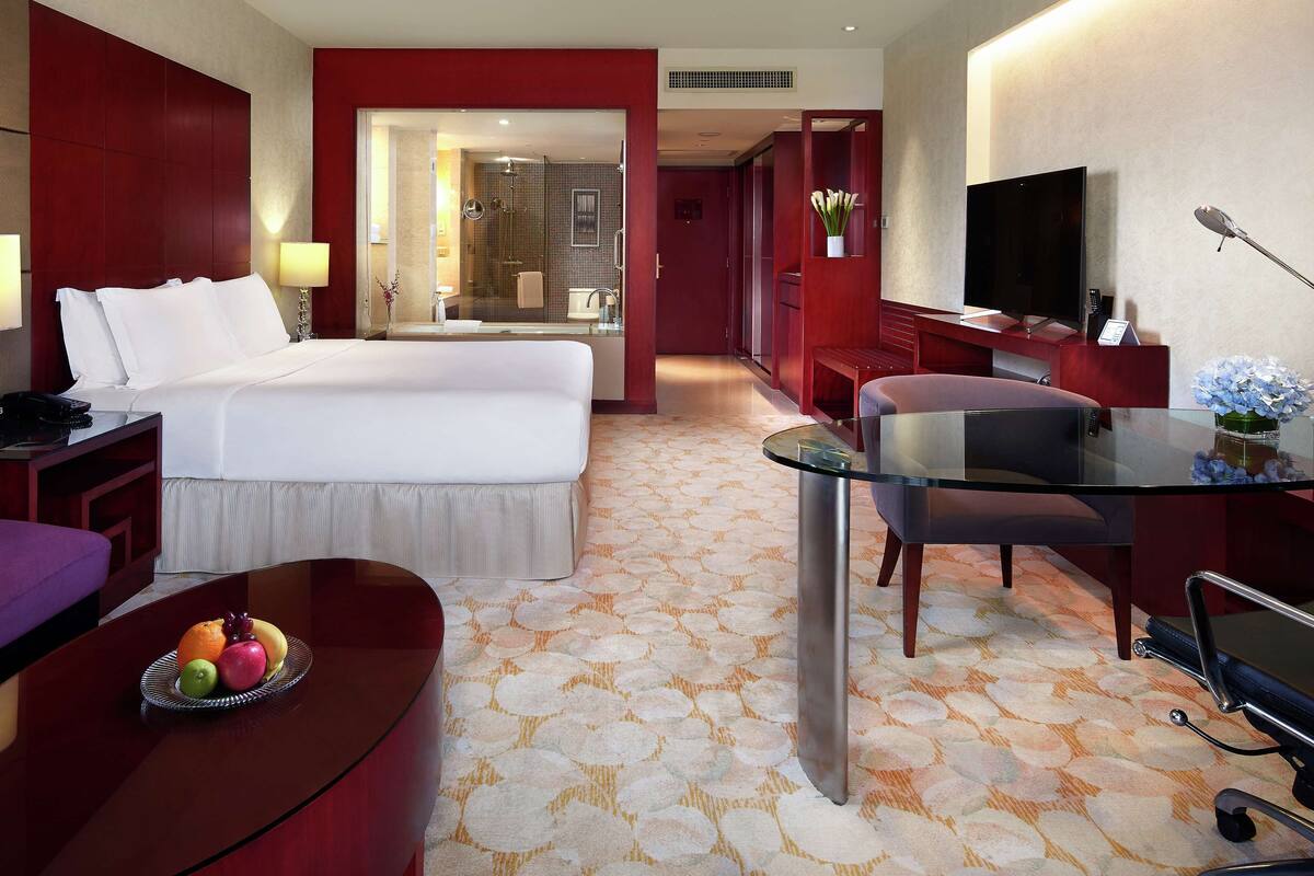Une chambre d’hôtel avec un lit, un bureau, une télévision, une table en verre et une salle de bain visible à travers une porte ouverte.