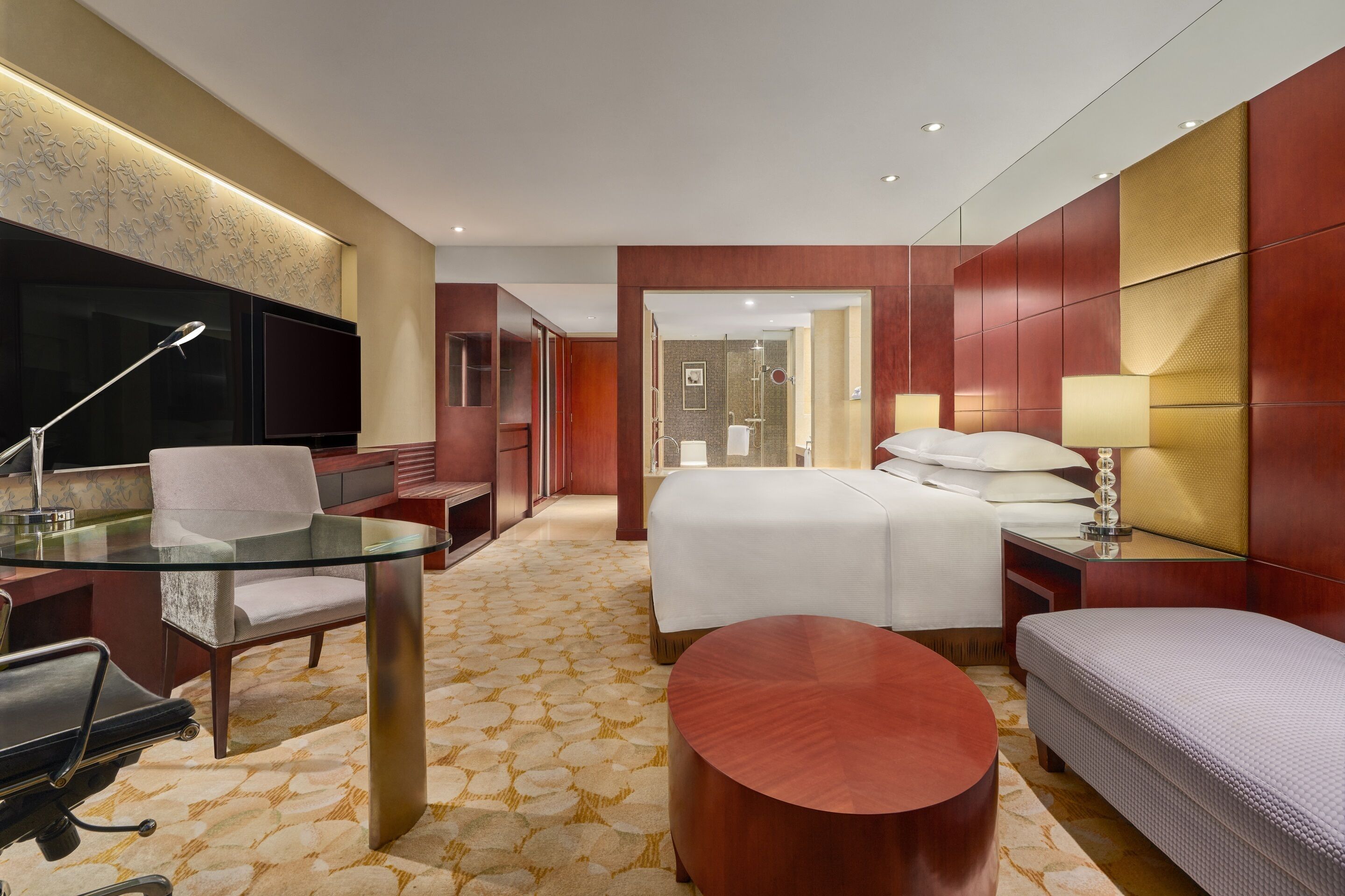A hotel room with a bed, a desk with a chair, a TV, and a bathroom visible through an open door.