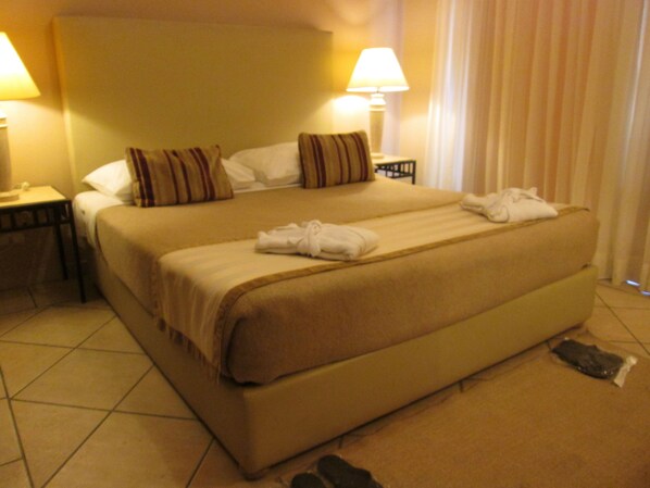 A large bed with a beige bedspread and pillows, two bedside lamps, and a pair of shoes on the floor.