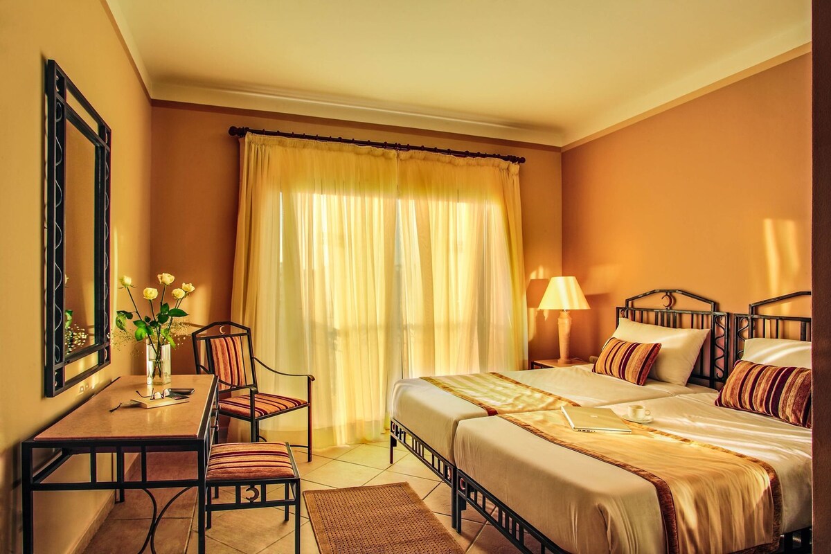 A hotel room with a bed, bedside table, chair, lamp, and a window with curtains.