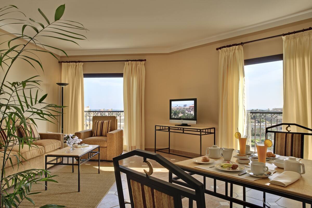 Habitación de hotel con balcón, televisor, mesa de comedor con desayuno servido y sofá.