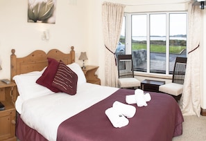 A neatly made bed with a wooden headboard, a red throw blanket, and white towels. There is a window with curtains, a chair, and a view of the outdoors. - Rusheen Bay House (Galway)