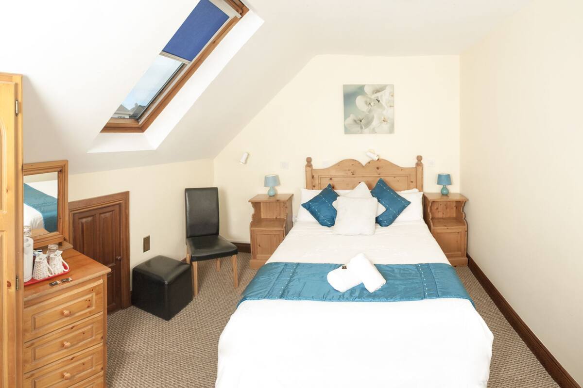 A bedroom with a skylight, a bed with blue bedding, a chair, a dresser, and a nightstand with a lamp.