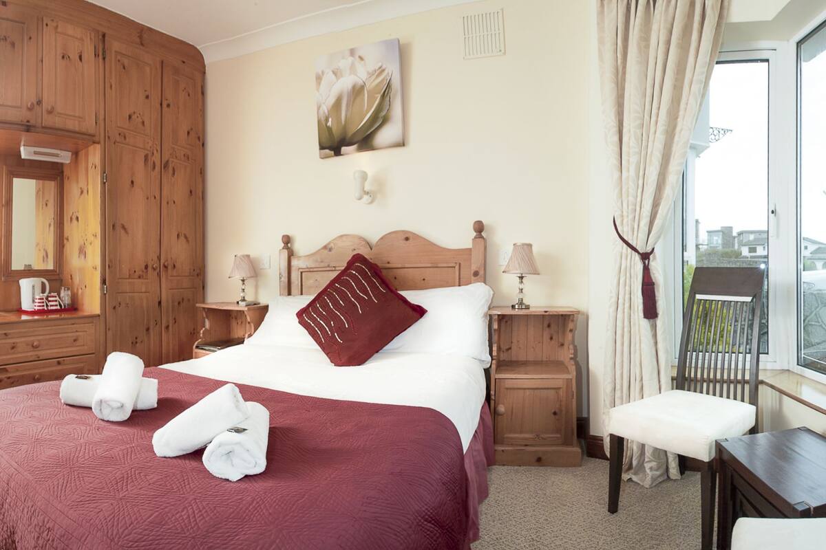 A hotel room with a wooden bed, two bedside tables with lamps, a window with curtains, a chair, and a view of the outside.