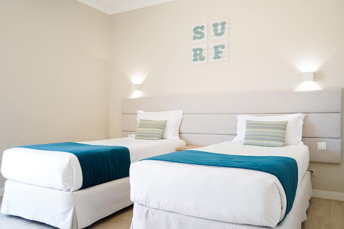 Ein Hotelzimmer mit zwei Einzelbetten, jeweils mit weißer Matratze und türkisblau gewählter Bettwäsche. An jeder Seite der Betten befinden sich Wandleuchten, und darüber hängt ein Schild mit der Aufschrift 'SURF'.