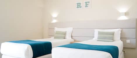 Ett hotellrum med två separata sängar, var och en med en vit madrass och turkosfärgat sänglinne. På vardera sidan av sängarna finns två väggmonterade lampor, och ovanför sängarna hänger en skylt med texten "SURF".