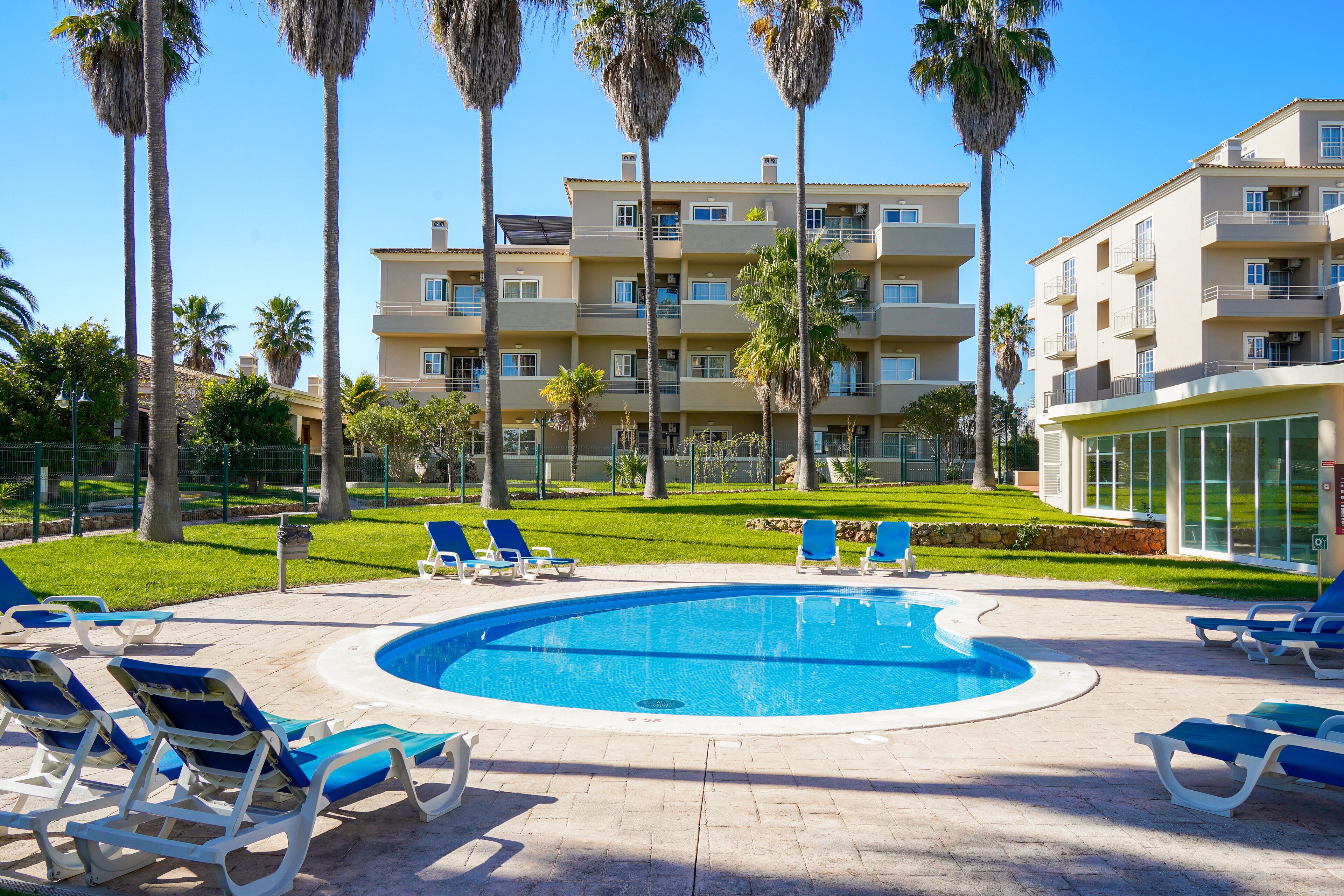 A swimming pool with lounge chairs, surrounded by palm trees and a multi-story building with balconies.