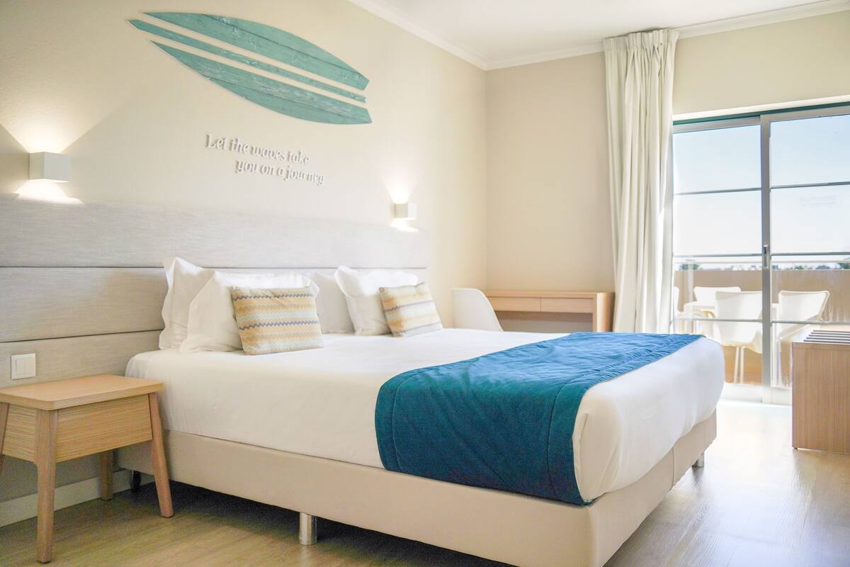 Ein Hotelzimmer mit einem großen Bett, einem Nachttisch, einer Wand mit Blattdekor und Text sowie einem Blick durch ein Fenster.