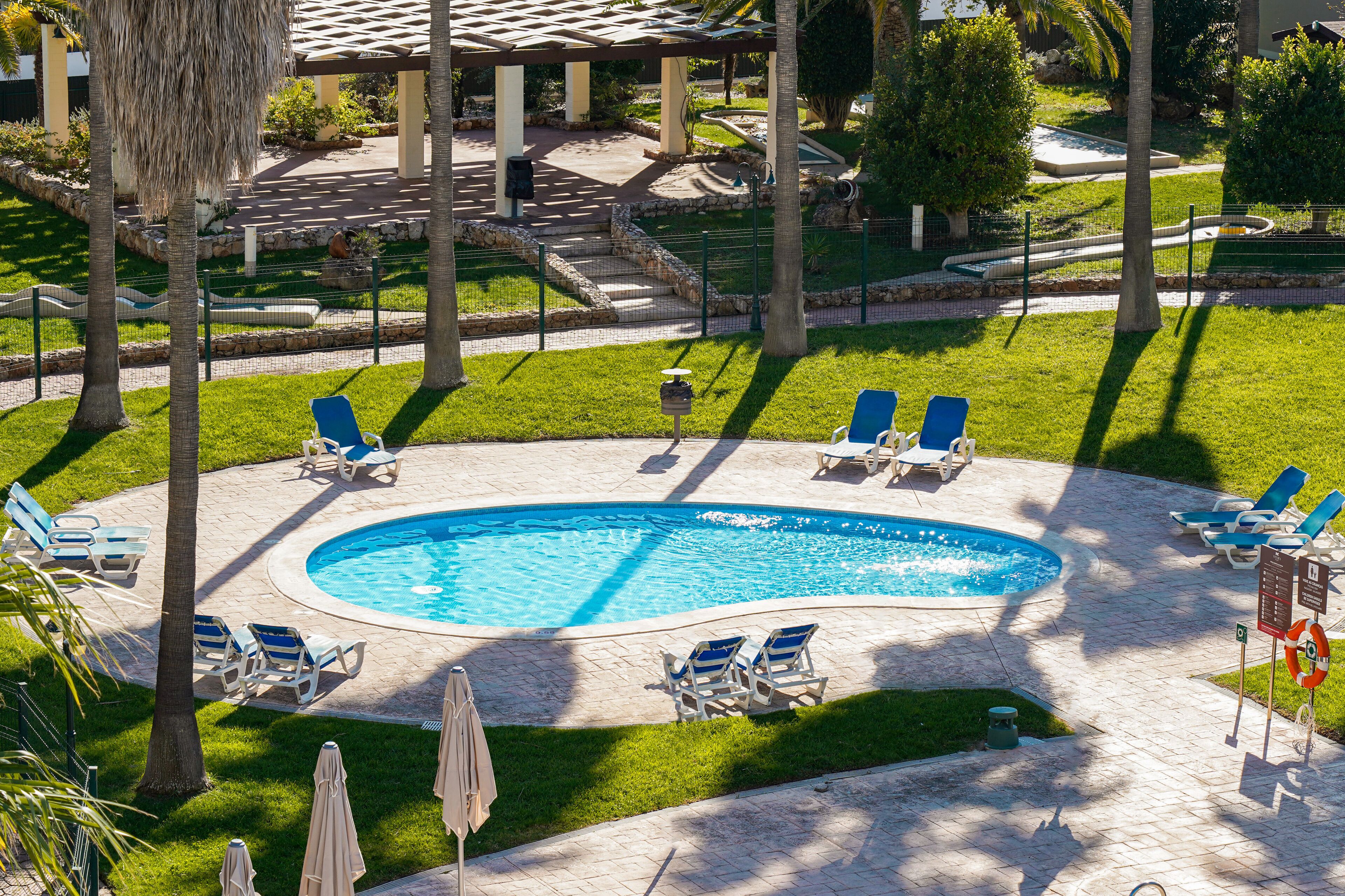 Ein Swimmingpool mit Liegestühlen und Sonnenschirmen, umgeben von einem angelegten Garten und einem überdachten Bereich mit Treppe.
