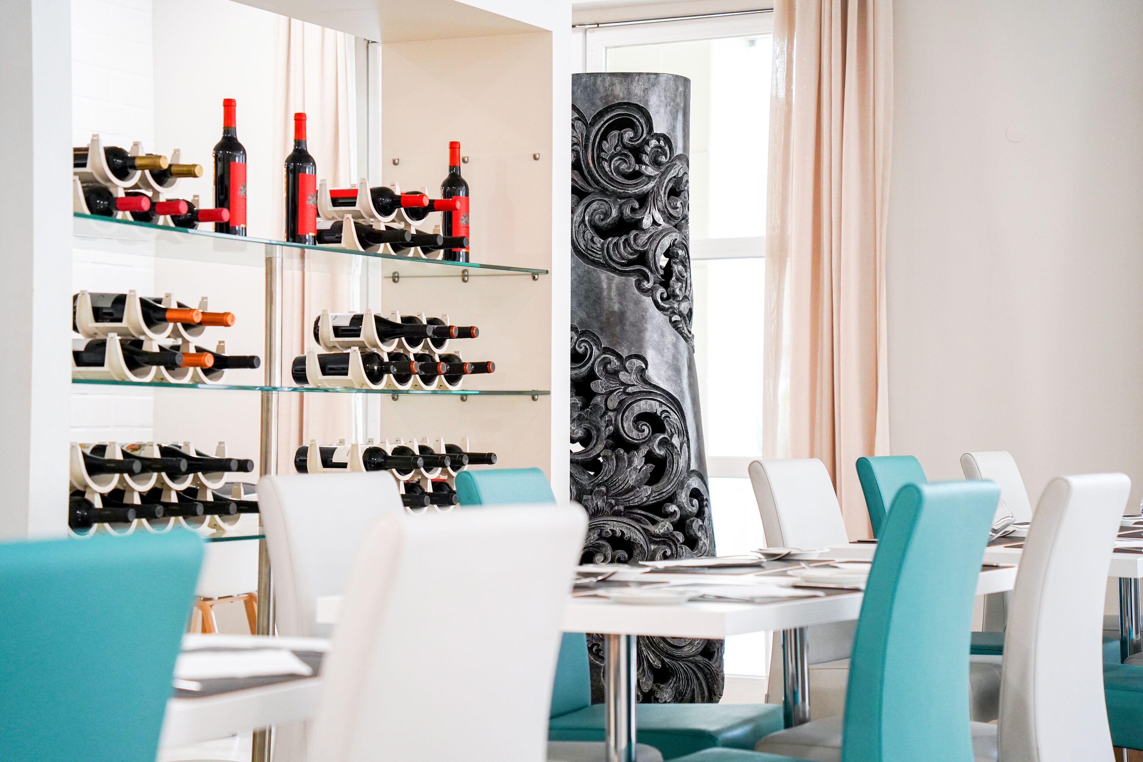 A dining area with glass shelves displaying wine bottles, teal chairs, and a decorative pillar.