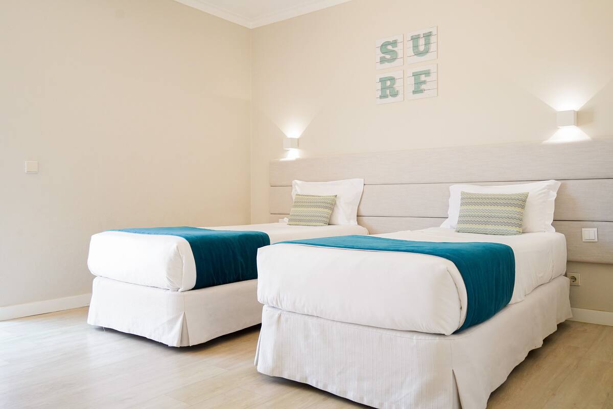 Ein Schlafzimmer mit zwei Betten, jeweils mit weißer Matratze und türkisblau gestreuter Bettwäsche. An den Wänden sind Leuchten angebracht, und ein Schild mit Buchstaben ist vorhanden.