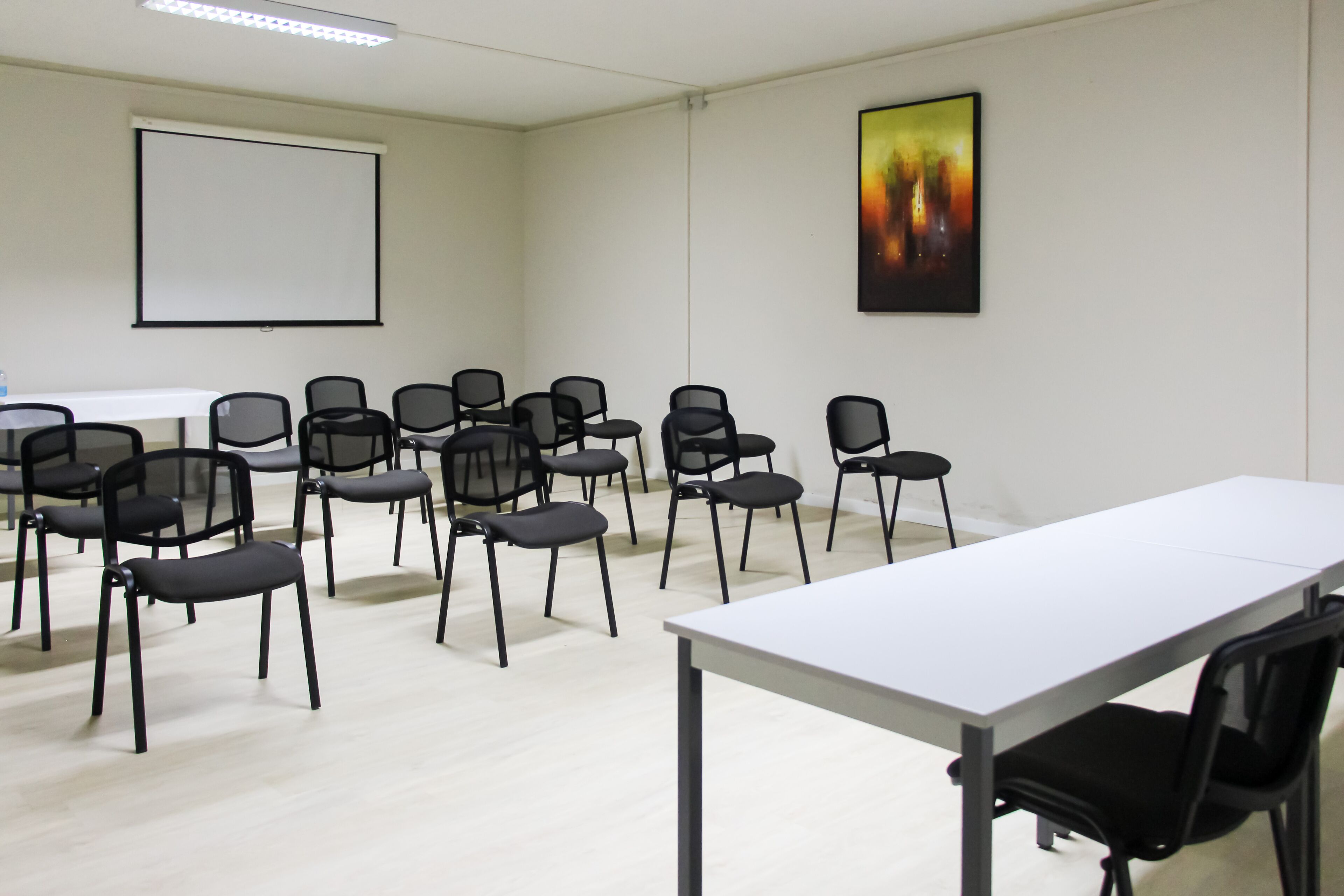 A conference room with a white table, black chairs, a projector screen, and a painting on the wall.
