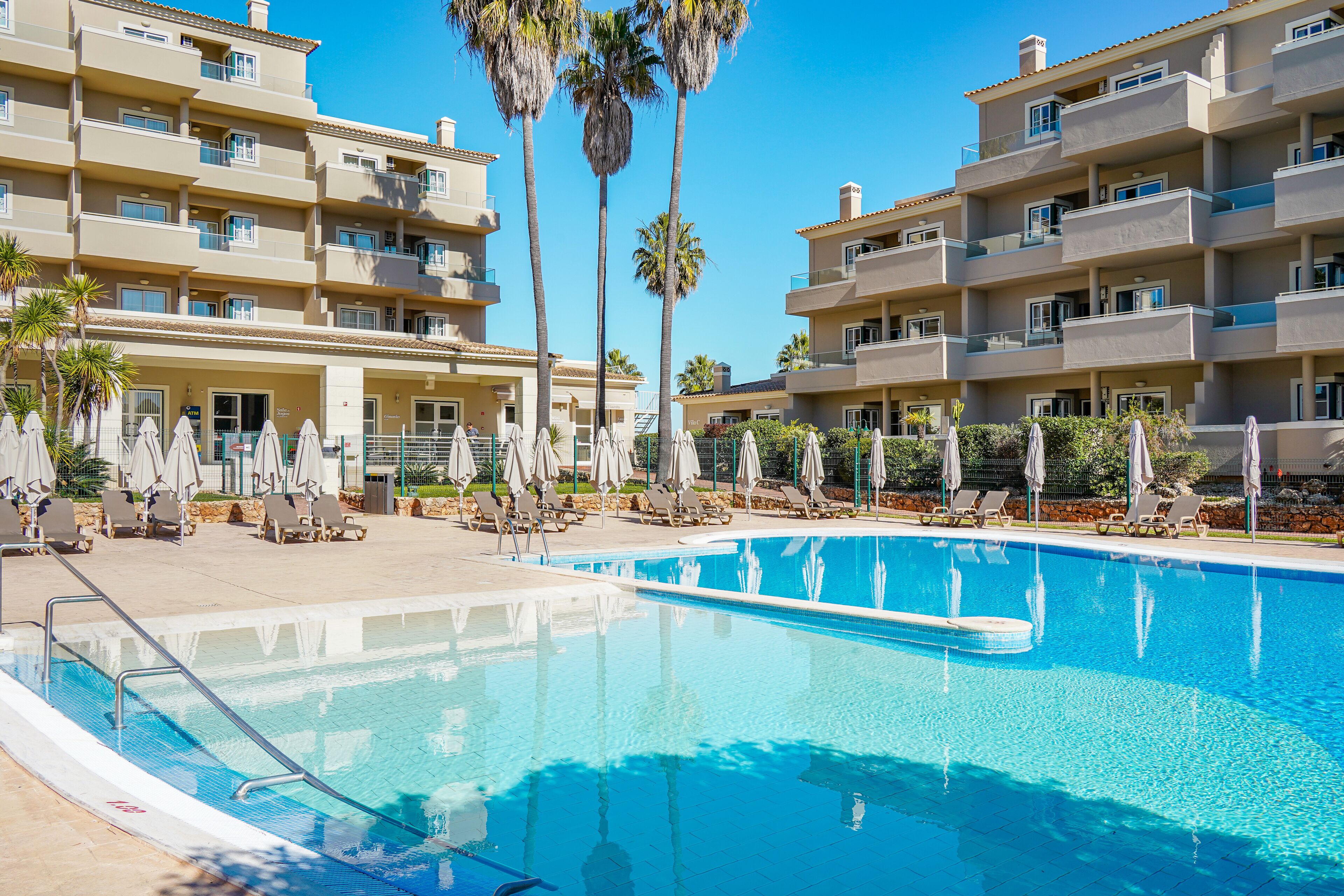 A swimming pool surrounded by lounge chairs and umbrellas, with multi-story buildings in the background.