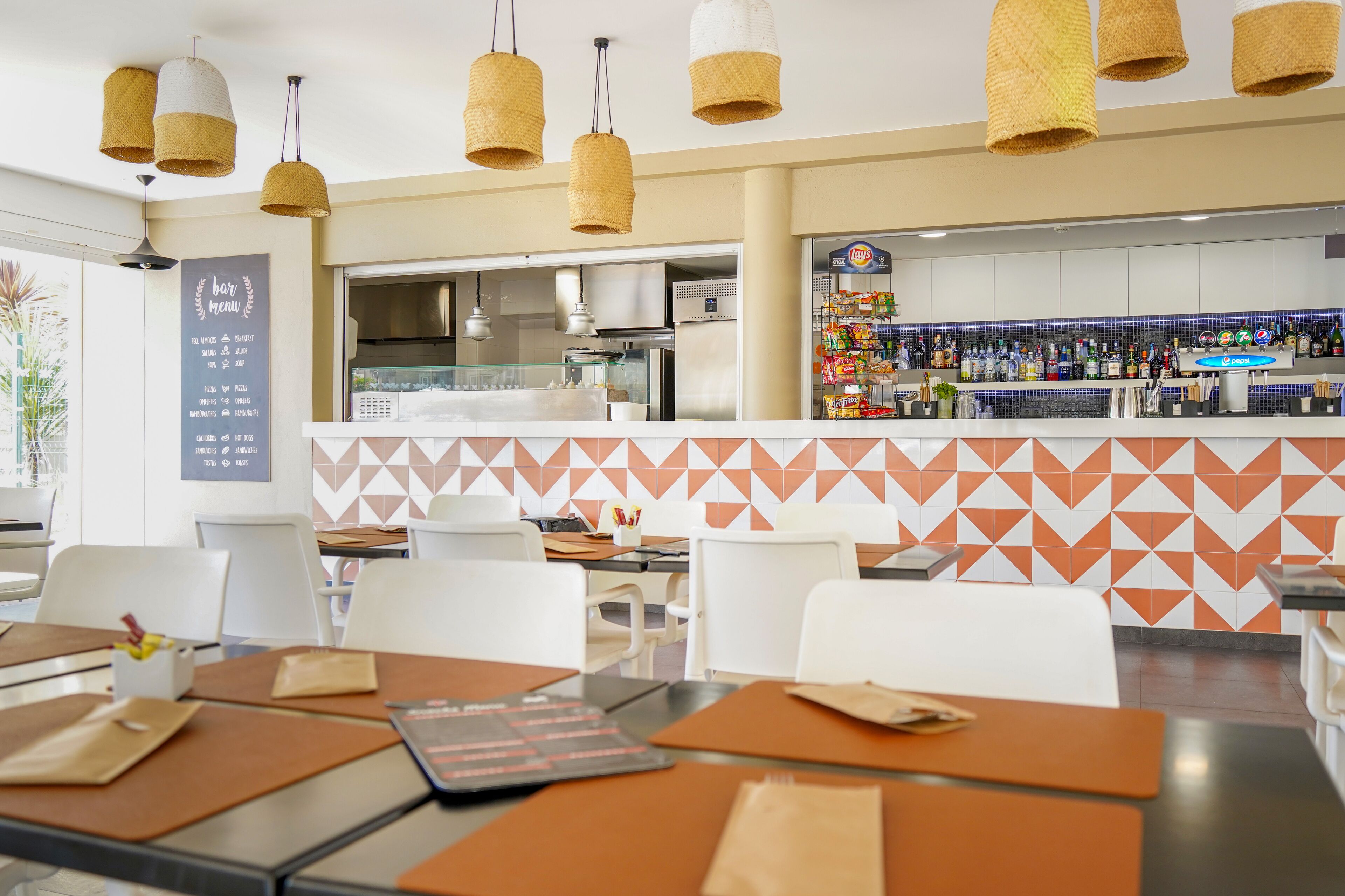 L'intérieur d'un restaurant, avec des tables, des chaises, un tableau affichant le menu, un espace cuisine et un bar proposant diverses boissons.