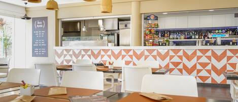 L'intérieur d'un restaurant, avec des tables, des chaises, un tableau affichant le menu, un espace cuisine et un bar proposant diverses boissons.