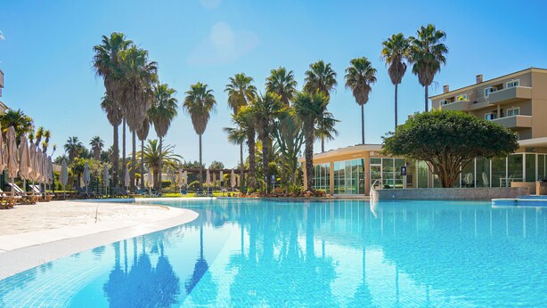 A swimming pool with clear blue water, surrounded by palm trees and lounge chairs. - Vitors Plaza (Portimão)
