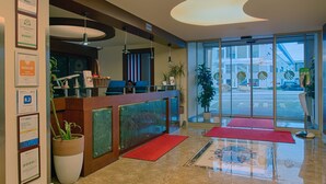 A hotel reception area with a modern design, including a reception desk, potted plants, and framed awards on the wall. - Airport Global Hotel  (Moerfelden-Walldorf)