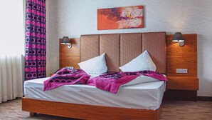 A hotel room with a wooden headboard, two bedside lamps, a bed with white linens, and a colorful blanket. - Airport Global Hotel  (Moerfelden-Walldorf)