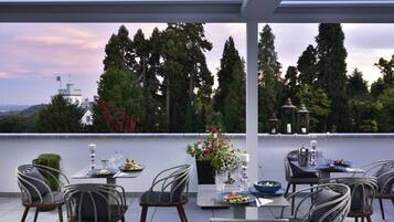 An outdoor dining area with tables and chairs, a view of trees and a distant building, and a lantern on the table.