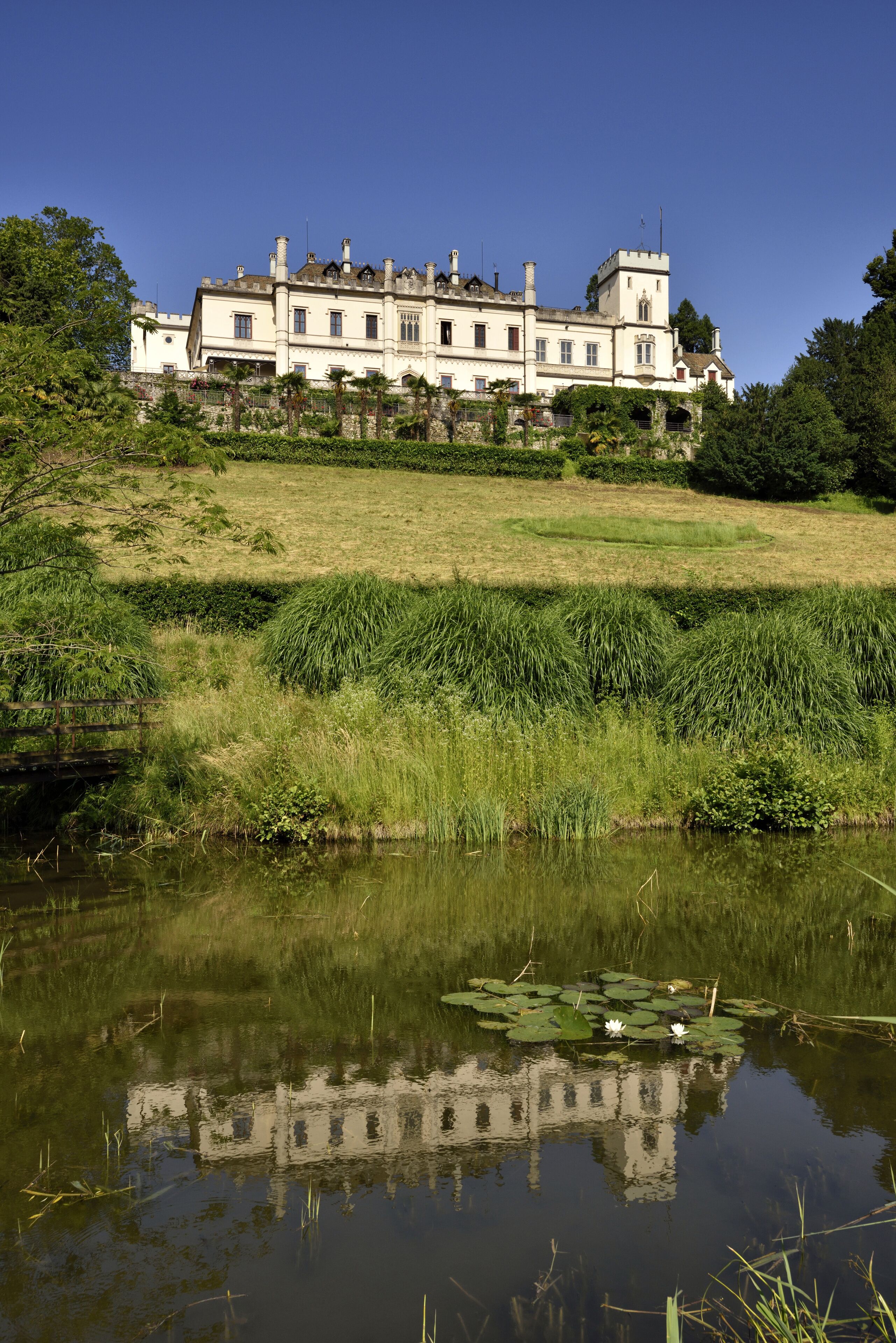 Un edificio grande e histórico con fachada simétrica, ubicado en una colina cubierta de césped con vista a una masa de agua tranquila con lirios.