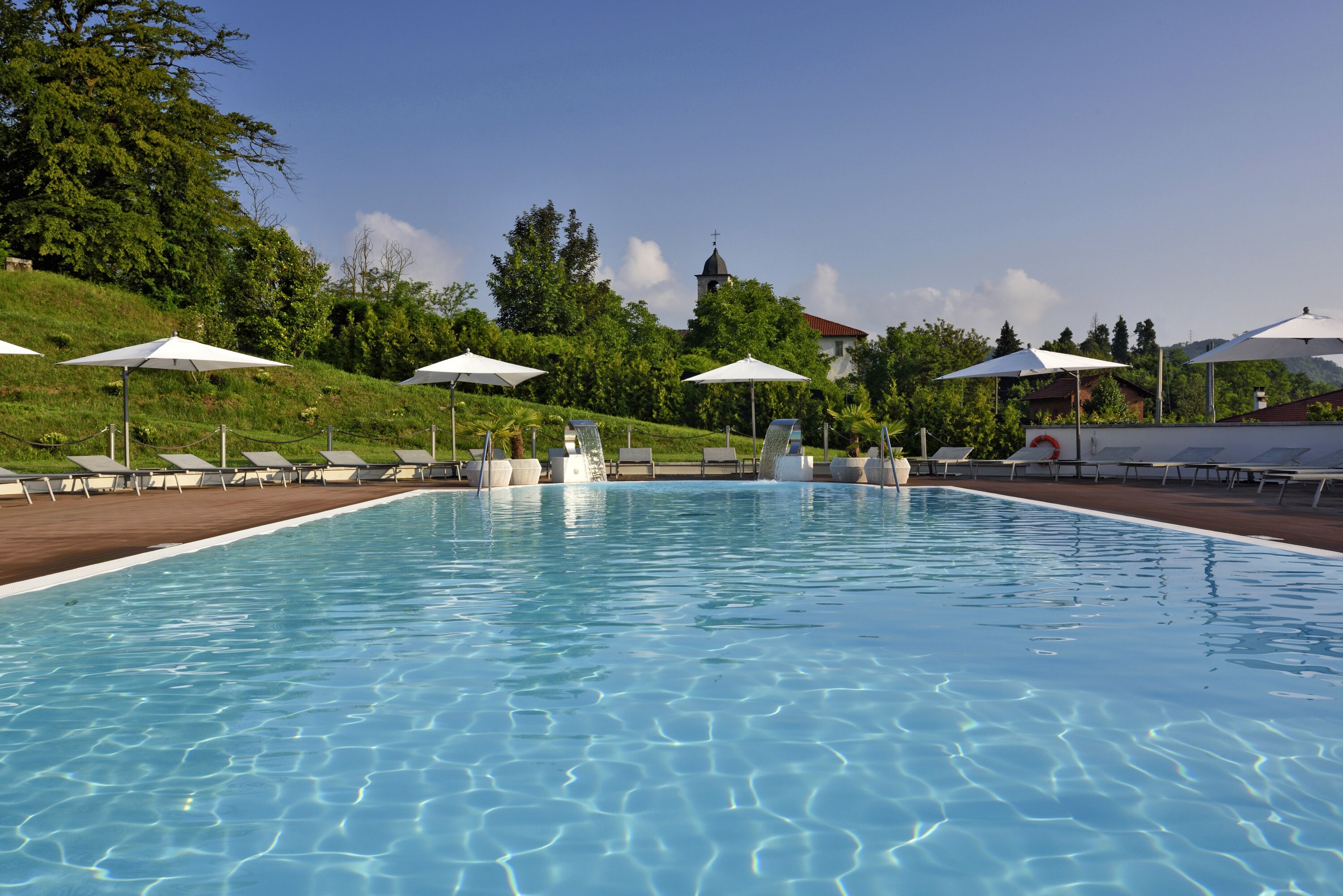 Une piscine entourée de chaises longues et de parasols, bordée de verdure et avec une colline en arrière-plan sur laquelle se trouve un bâtiment.