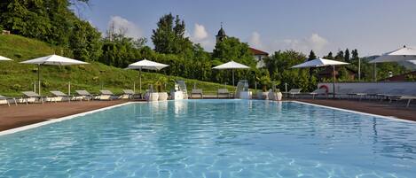 Une piscine entourée de chaises longues et de parasols, bordée de verdure et avec une colline en arrière-plan sur laquelle se trouve un bâtiment.